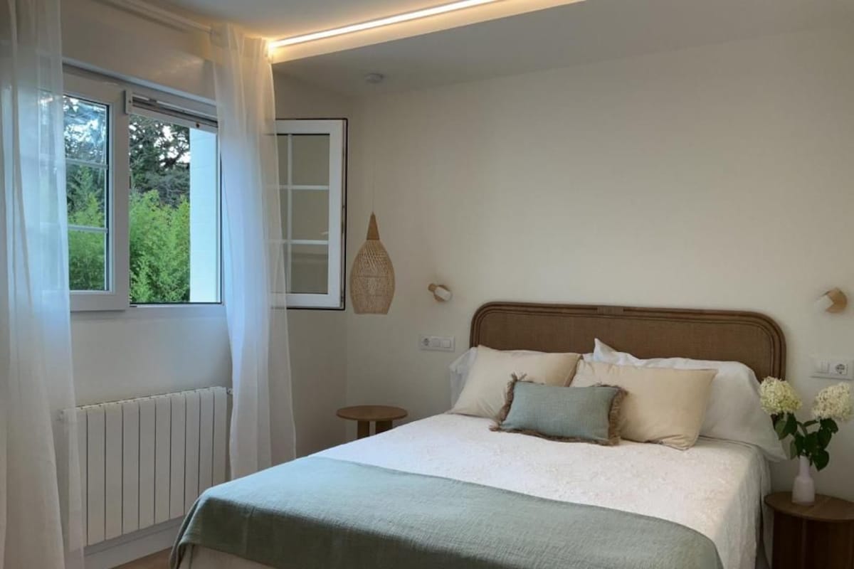 Ein Schlafzimmer mit Bett, Fenster mit Vorhängen, Nachttisch mit Vase und einer Wandleuchte.