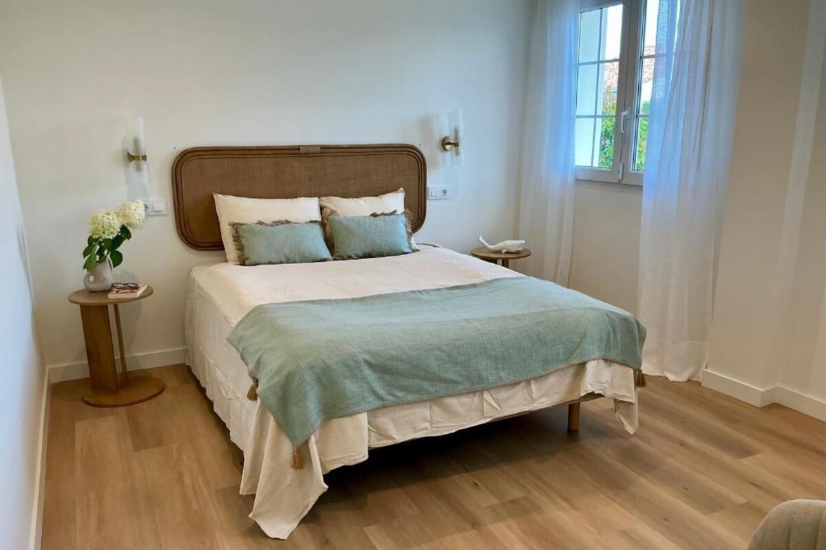 Ein Schlafzimmer mit einem großen Bett, Nachttischen, einem Fenster mit Vorhängen und Blick auf Grünfläche.