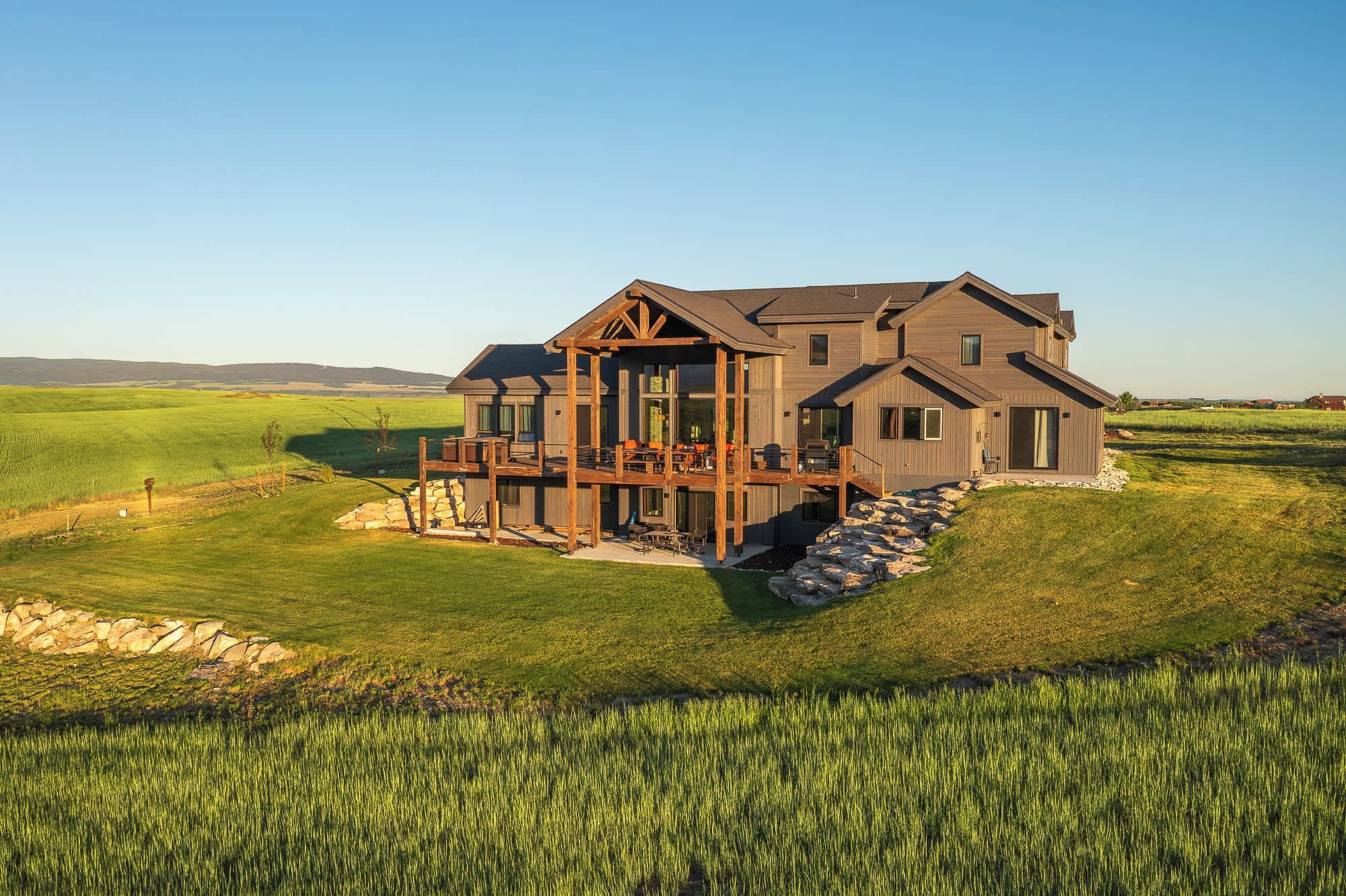 A large, two-story wooden house with a deck and multiple windows, surrounded by a grassy field and a stone wall.