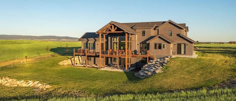 A large, two-story wooden house with a deck and multiple windows, surrounded by a grassy field and a stone wall.