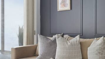Una sala de estar con un sofá color beige, almohadas grises y blancas, una mesa de centro de vidrio con un libro y un globo terráqueo, y un cuadro abstracto enmarcado en la pared.