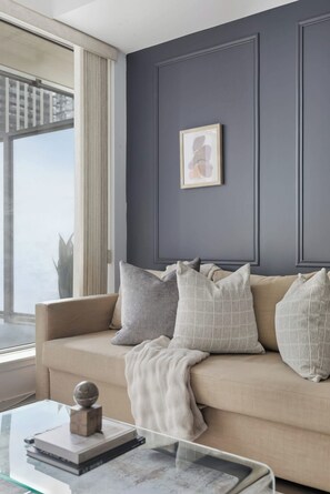 Una sala de estar con un sofá color beige, almohadas grises y blancas, una mesa de centro de vidrio con un libro y un globo terráqueo, y un cuadro abstracto enmarcado en la pared.