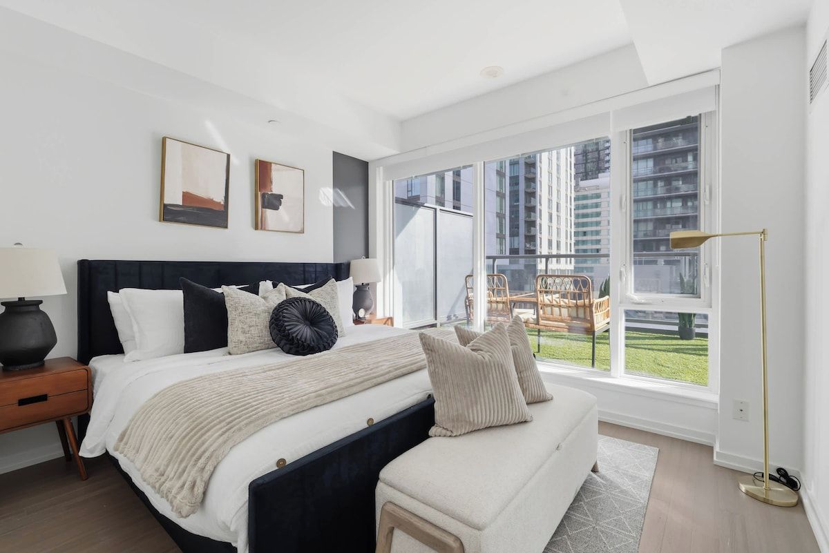 Un dormitorio moderno con una cama grande, mesitas de noche, un banco y vistas a un paisaje urbano a través de amplios ventanales.