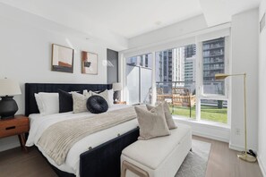 Un dormitorio moderno con una cama grande, mesitas de noche, un banco y vistas a un paisaje urbano a través de amplios ventanales.