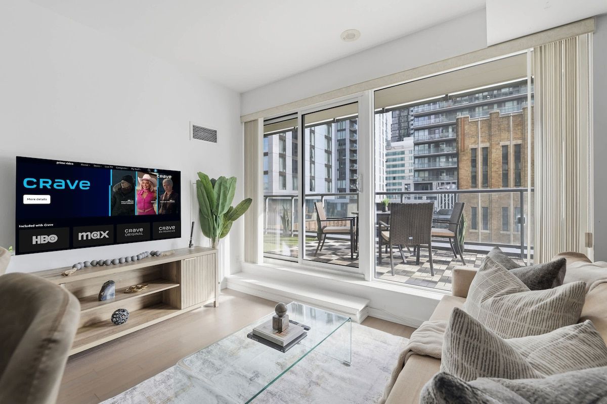 Una sala de estar moderna con un televisor grande mostrando un programa, un balcón con vistas a edificios y una zona de estar acogedora.