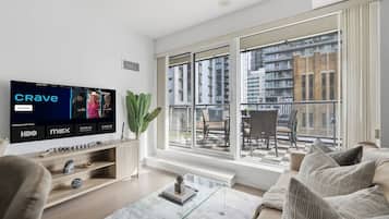 Una sala de estar moderna con un televisor grande mostrando un programa, un balcón con vistas a edificios y una zona de estar acogedora.