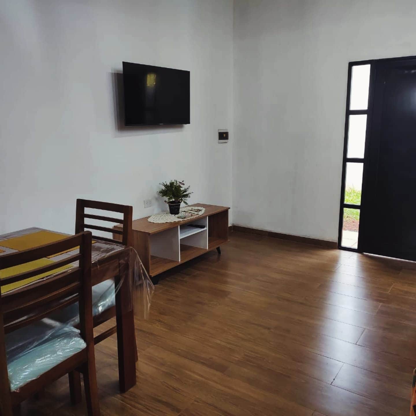 Une pièce avec un plancher en bois, une table à manger avec des chaises, une petite table sur laquelle est posée une plante, un téléviseur accroché au mur et une porte donnant sur l’extérieur.