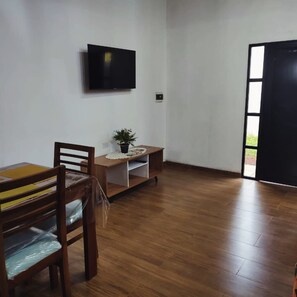 A room with a wooden floor, a dining table with chairs, a small table with a plant, a television on the wall, and a door leading outside.