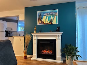 A living room with a fireplace, a painting, and a kitchen in the background.