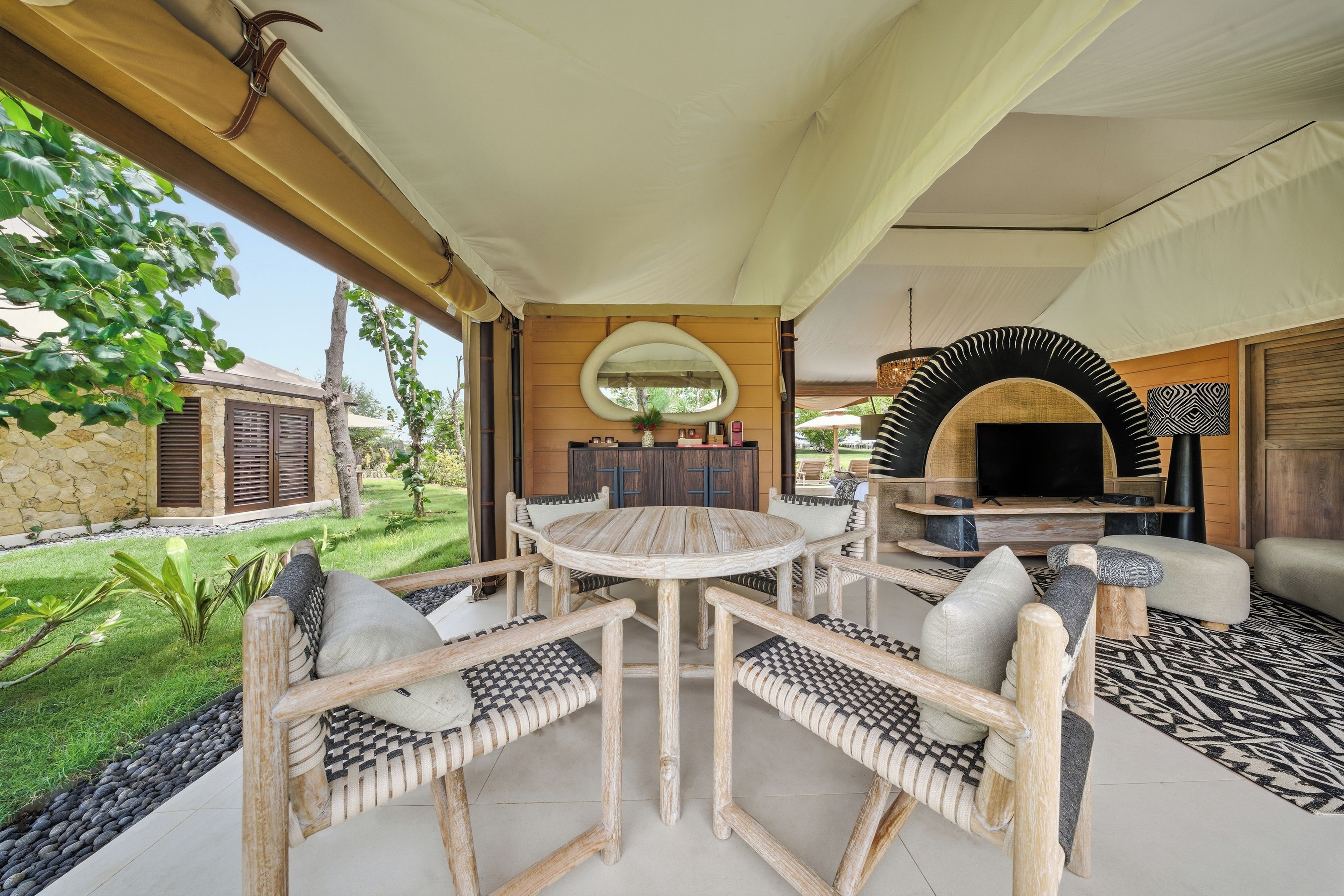 An outdoor living area with a round wooden table, cushioned chairs, a stone wall, and a view of a garden.