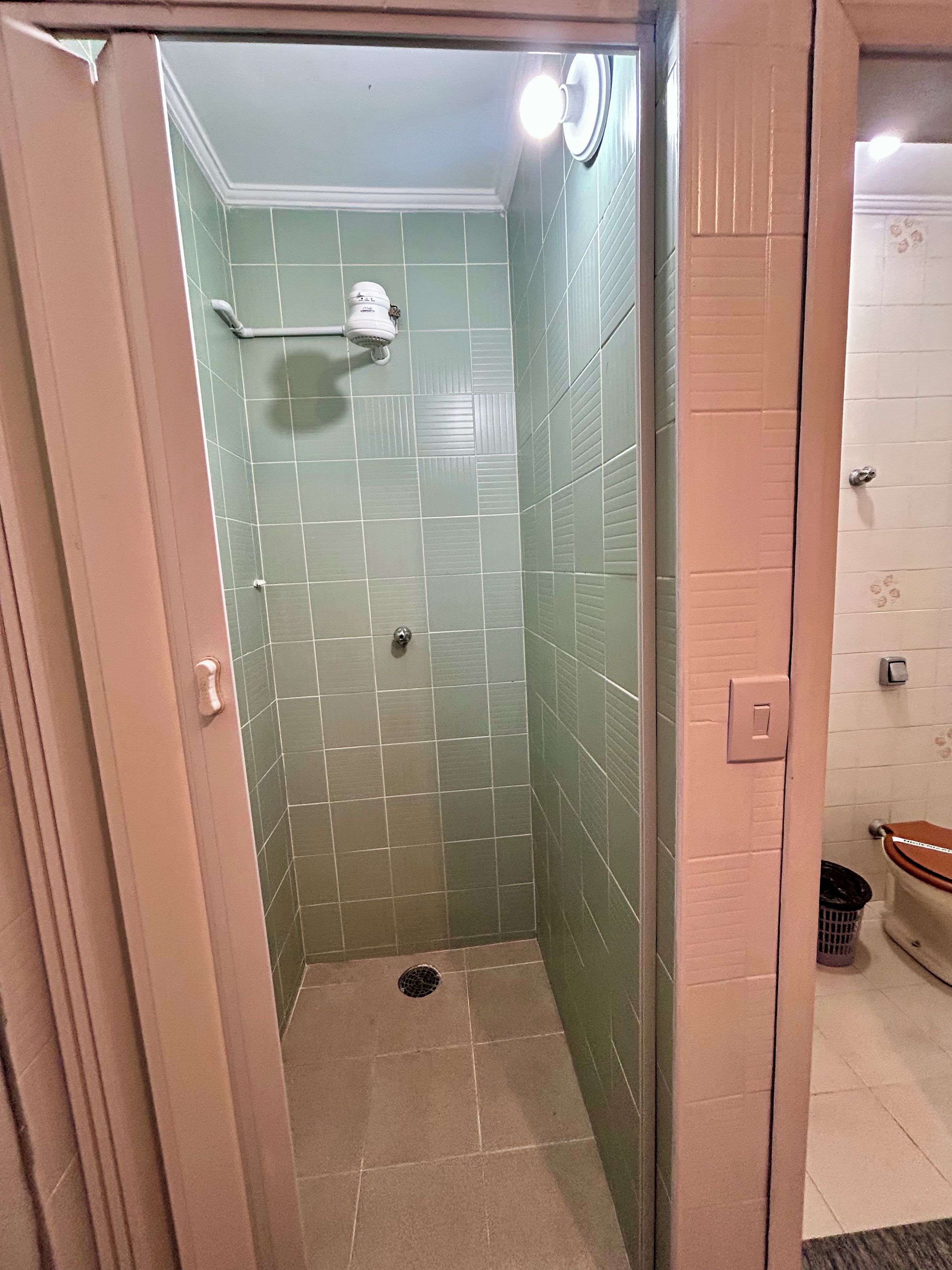 Um chuveiro pequeno e fechado, com azulejos verdes e uma luminária branca no teto.