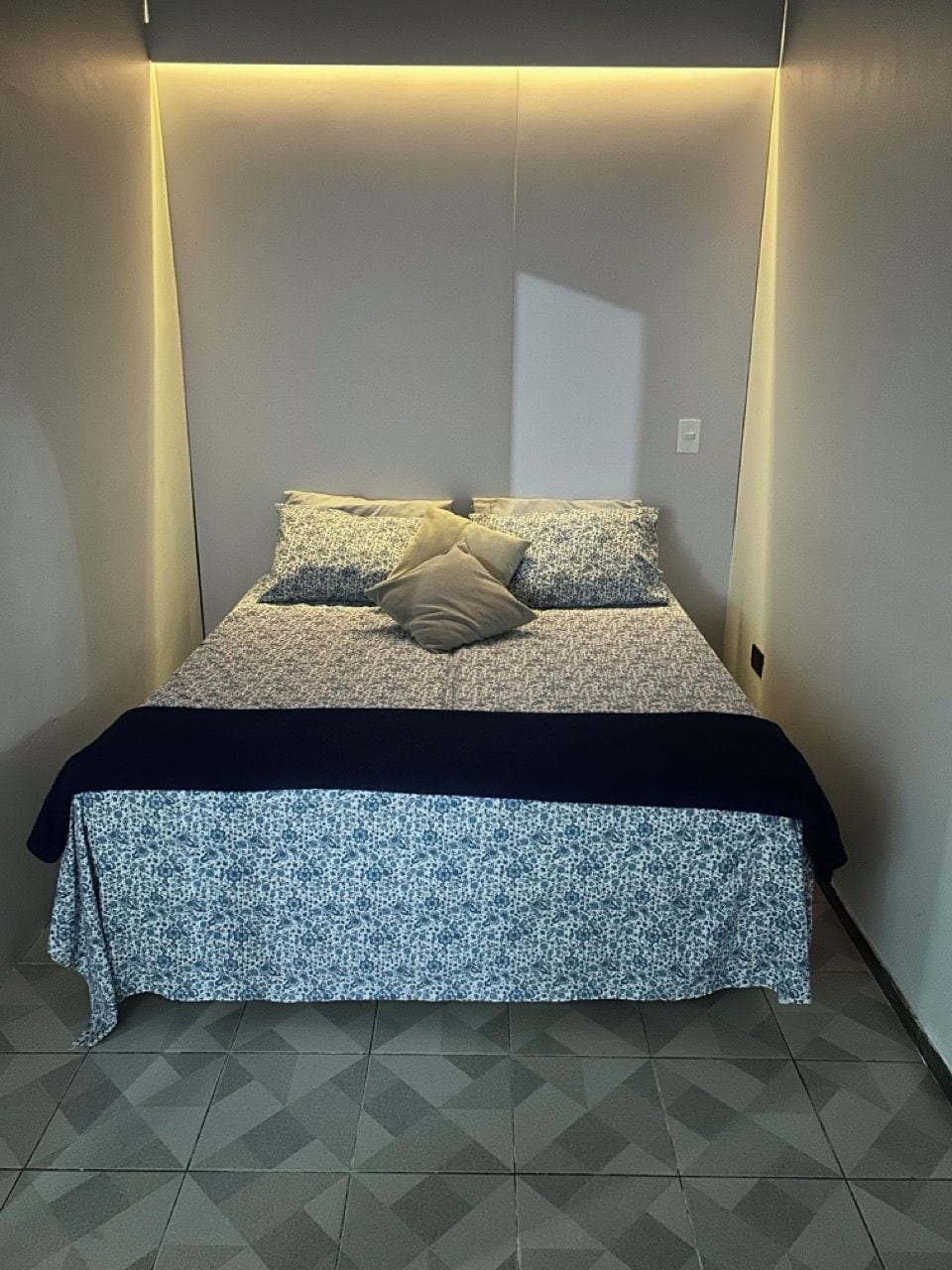 A bed with a patterned bedspread and pillows, a dark-colored blanket, and a geometric tile floor.