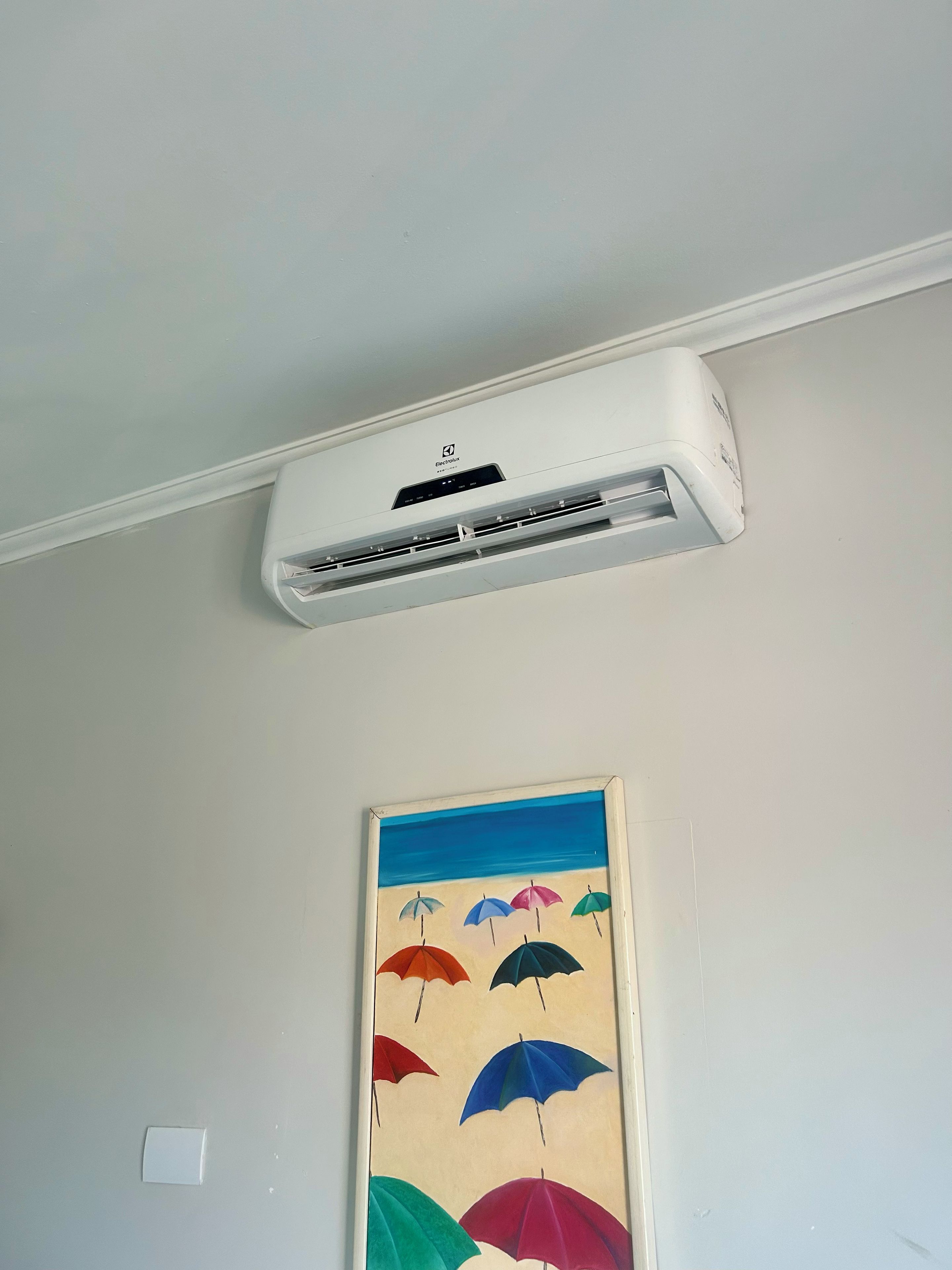 A white air conditioning unit mounted on a wall with a painting of colorful umbrellas below it.