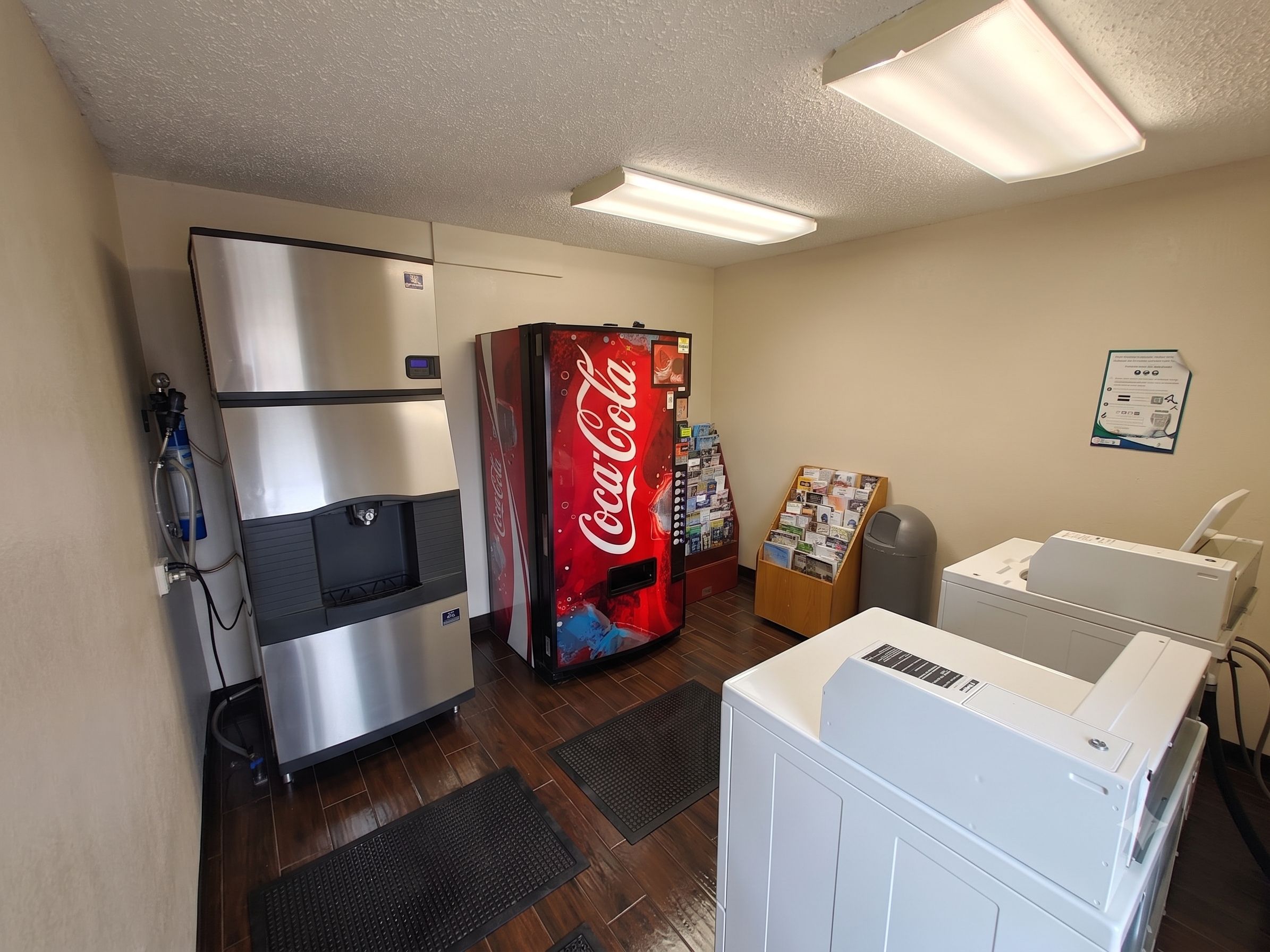 A room with a refrigerator, a Coca-Cola vending machine, a bulletin board, a trash bin, and a dryer.