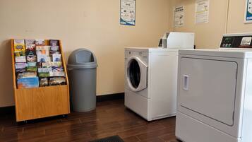A laundry area with a wooden rack of brochures, a gray trash bin, a front-loading washing machine, and a stacked dryer.