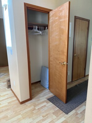 Una habitación con puerta de madera, un armario con ropa colgada, un cojín azul y una alfombra estampada.