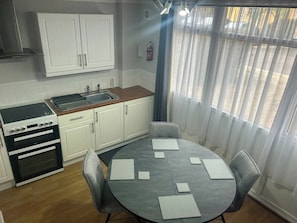 A kitchen with white cabinets, a stove, and a sink. There is a round table with chairs and papers on it. - 2 Bedroom Villa, Sleeps 5, Wifi, Parking, Beach (Dawlish)