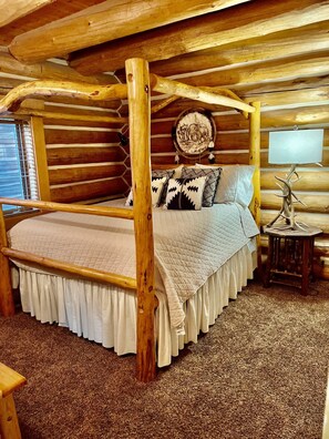 A log cabin bedroom with a four-poster bed, a round wall hanging, and a small table with a decorative item. - A Sundance Original • Historic Log Cabin (Sundance)