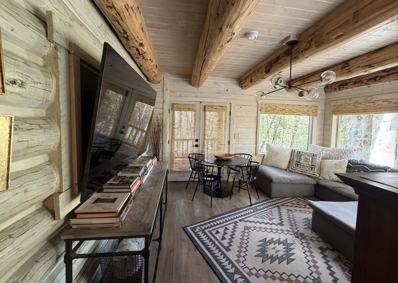 A cozy cabin interior with a wooden interior, a flat-screen TV on a stand, a sofa, a dining table with chairs, and a patterned rug.