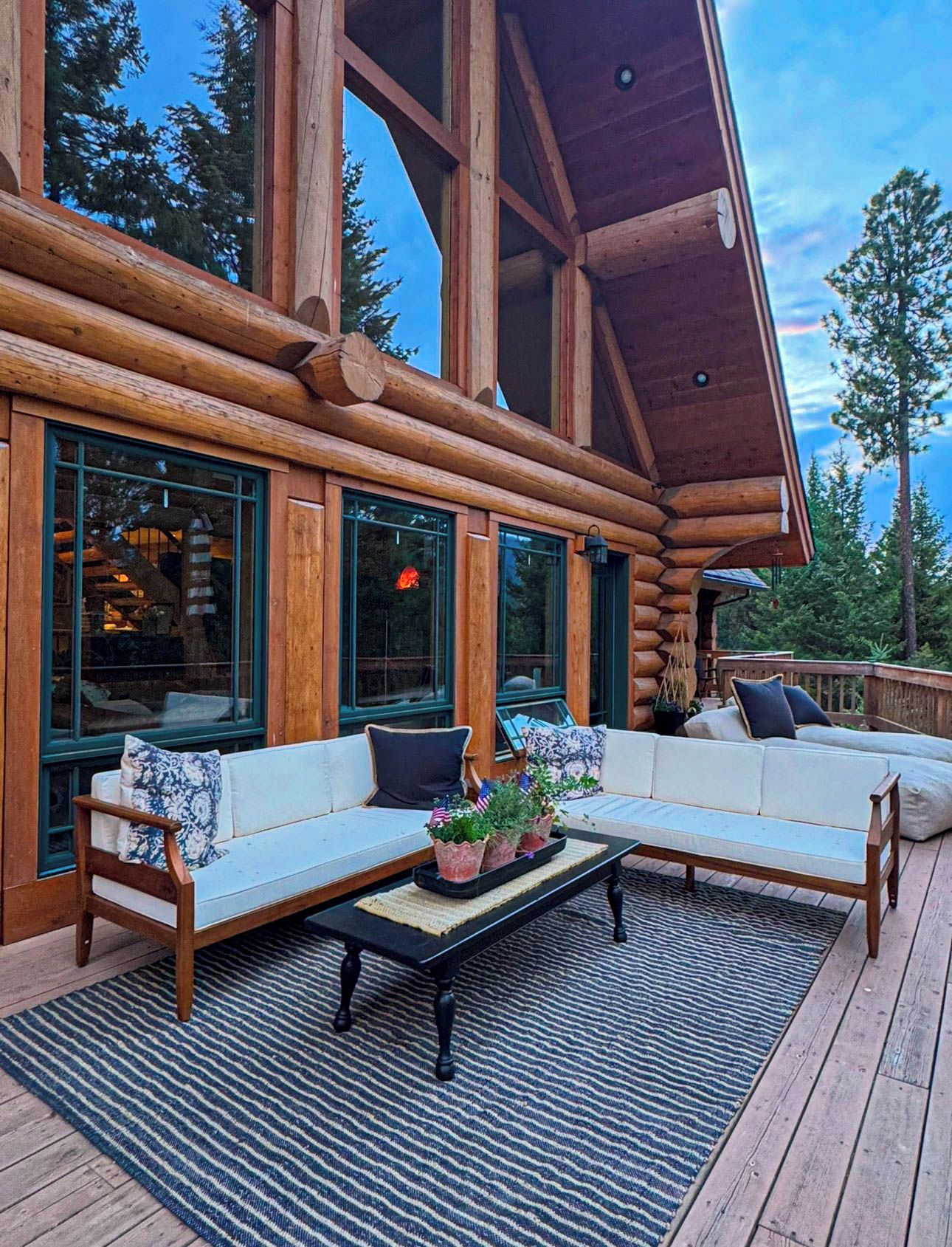 A log cabin with a large deck, outdoor seating, and a view of the surrounding forest.