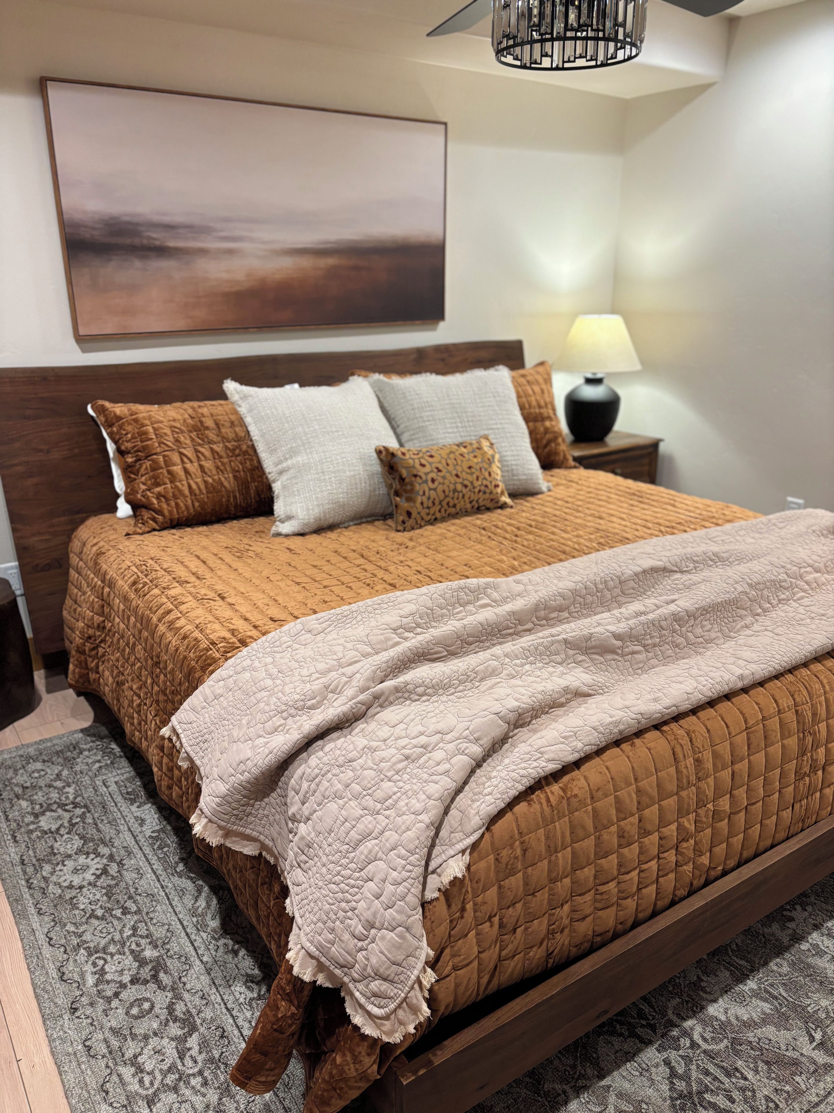 Un lit bien fait, recouvert d’une couverture marron, un tapis gris à motifs, une tête de lit en bois, une lampe de chevet et une œuvre d’art accrochée au mur.
