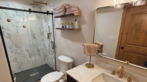 Ein modernes Badezimmer mit einer Duschkabine aus Glas, einer Toilette, einem Waschbecken mit goldfarbenen Armaturen und einem Schminktisch mit Spiegel.