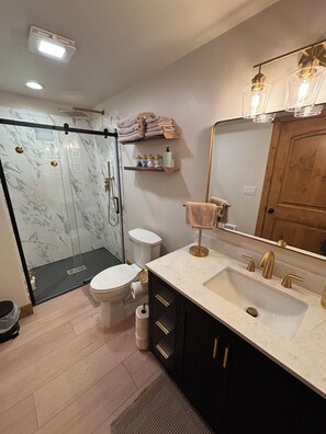Et moderne badeværelse med brusekabine i glas, toilet, vask med guldfarvede armaturer og et spejl med underskab.