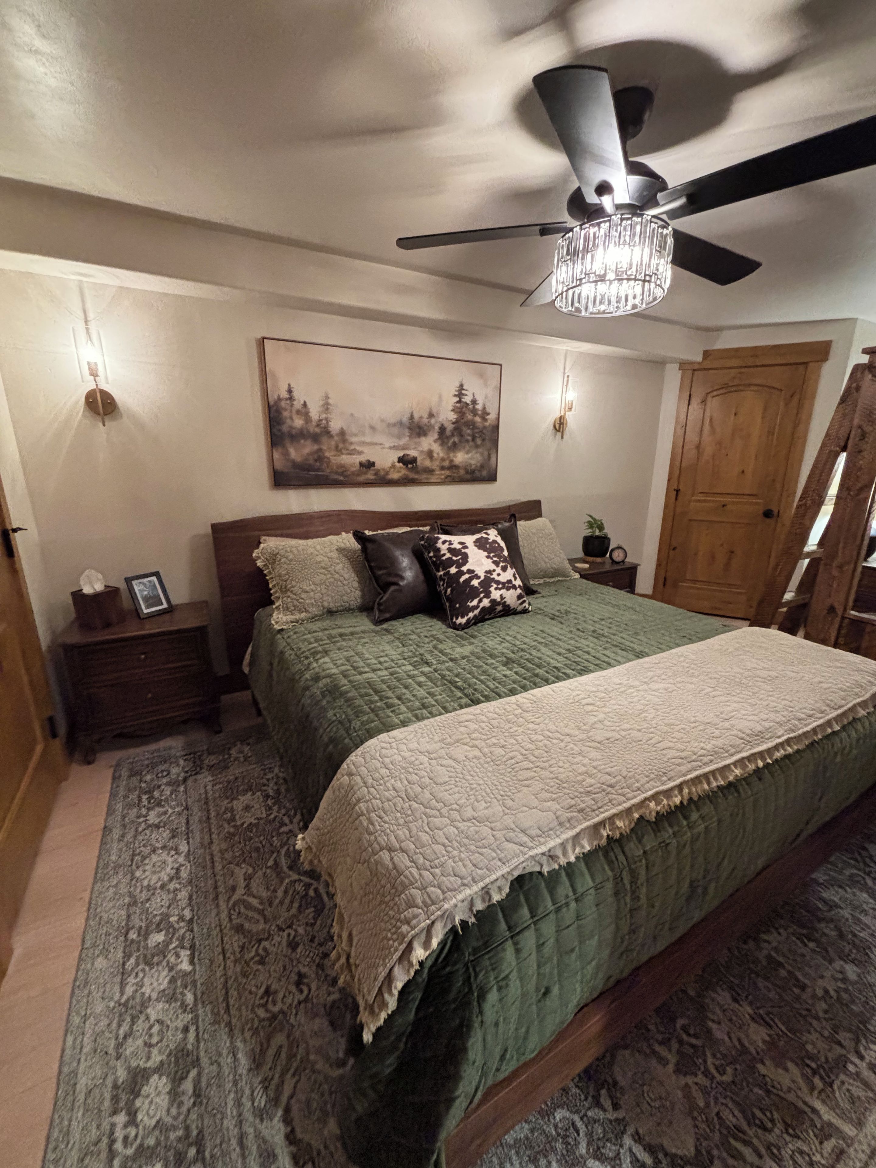 A bedroom with a bed, a ceiling fan, a painting, a wooden door, and a nightstand.