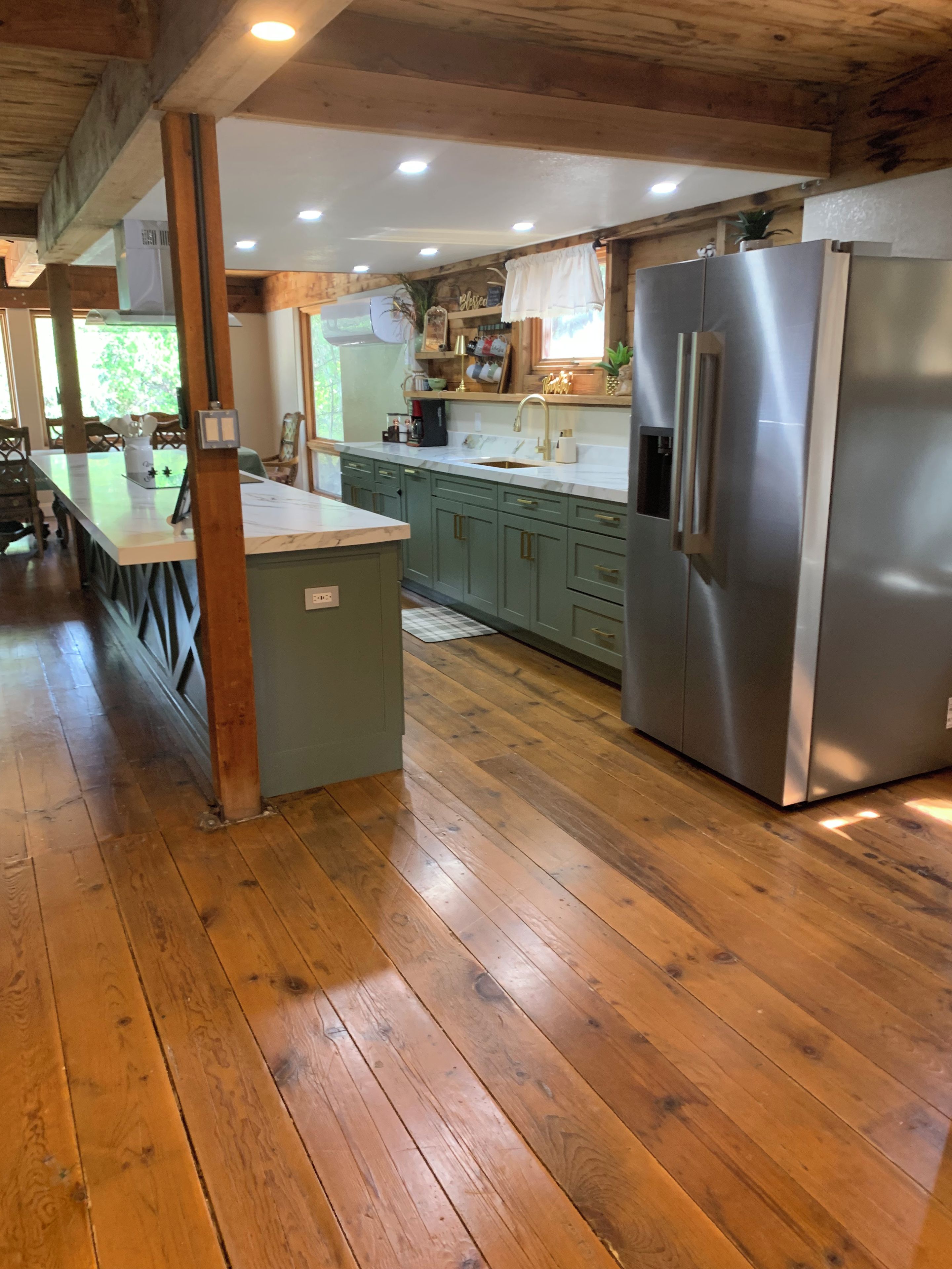 A kitchen with wooden floors, green cabinets, a stainless steel refrigerator, and a marble countertop.