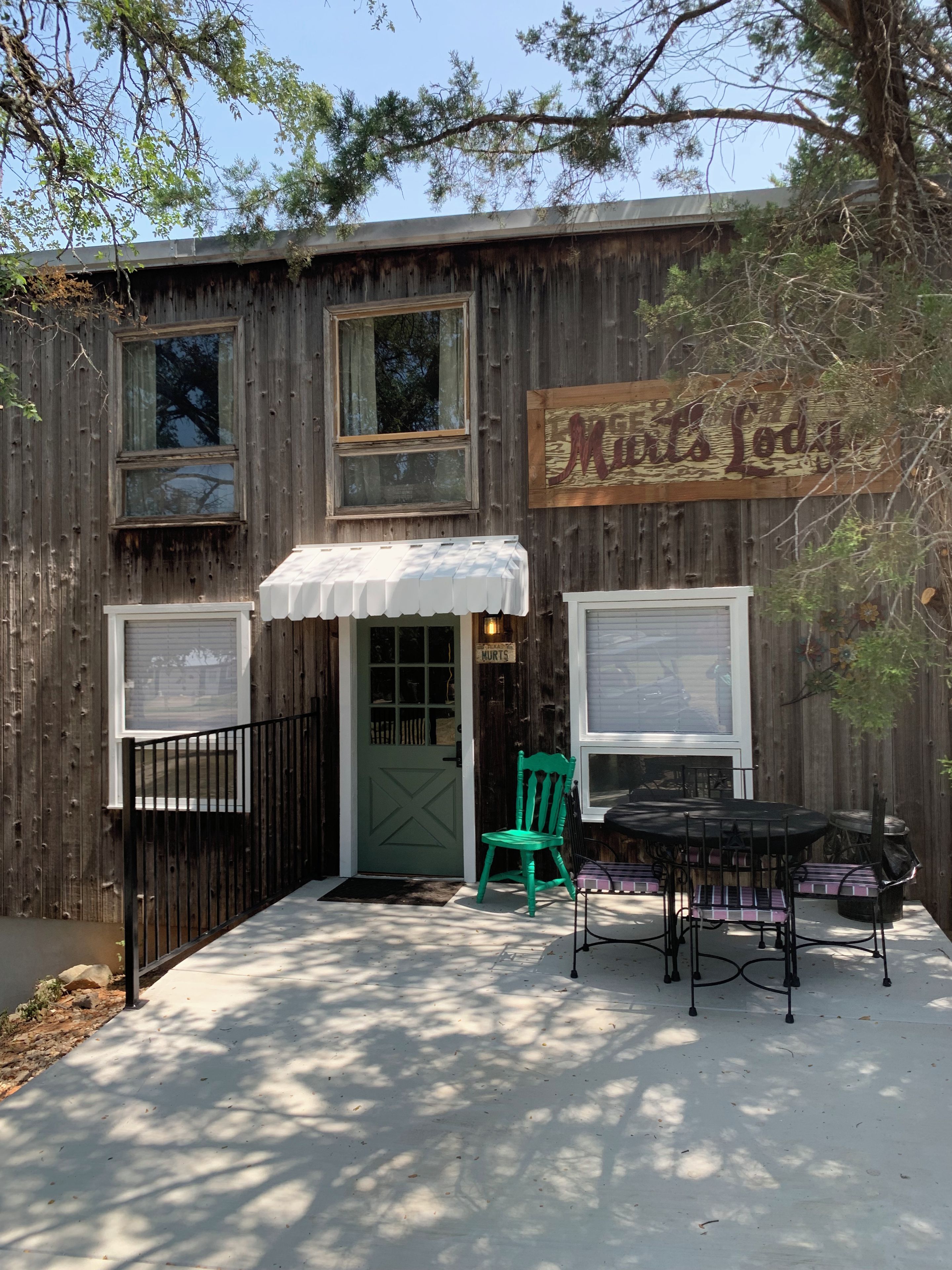 Un edificio de madera rústico con un letrero que dice 'Mats Lodge'. Hay una silla verde y una mesa con sillas afuera.