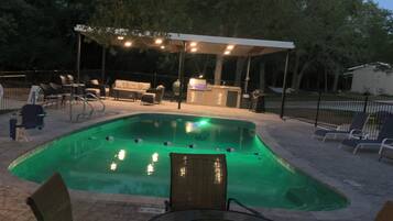 Zona de piscina exterior con área de estar cubierta, sillas de descanso y una estación de barbacoa.