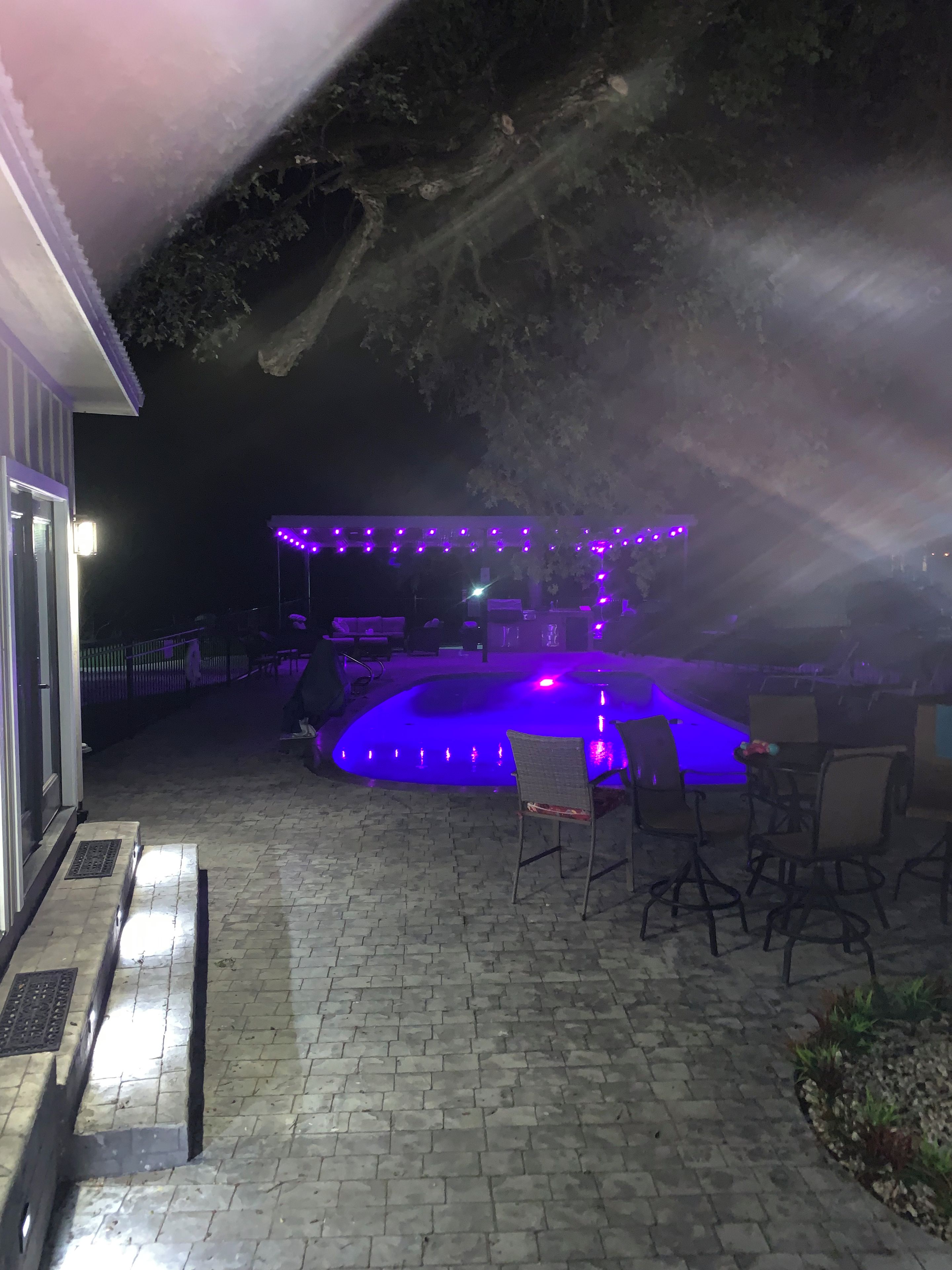 Un espace extérieur aménagé en terrasse, avec une piscine, entouré de chaises et de tables, et illuminé par des lumières violettes.