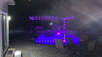 Un espace extérieur aménagé en terrasse, avec une piscine, entouré de chaises et de tables, et illuminé par des lumières violettes.