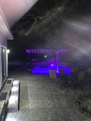 Zona de pati exterior amb una piscina, envoltada de cadires i taules, il·luminada amb llums morades.