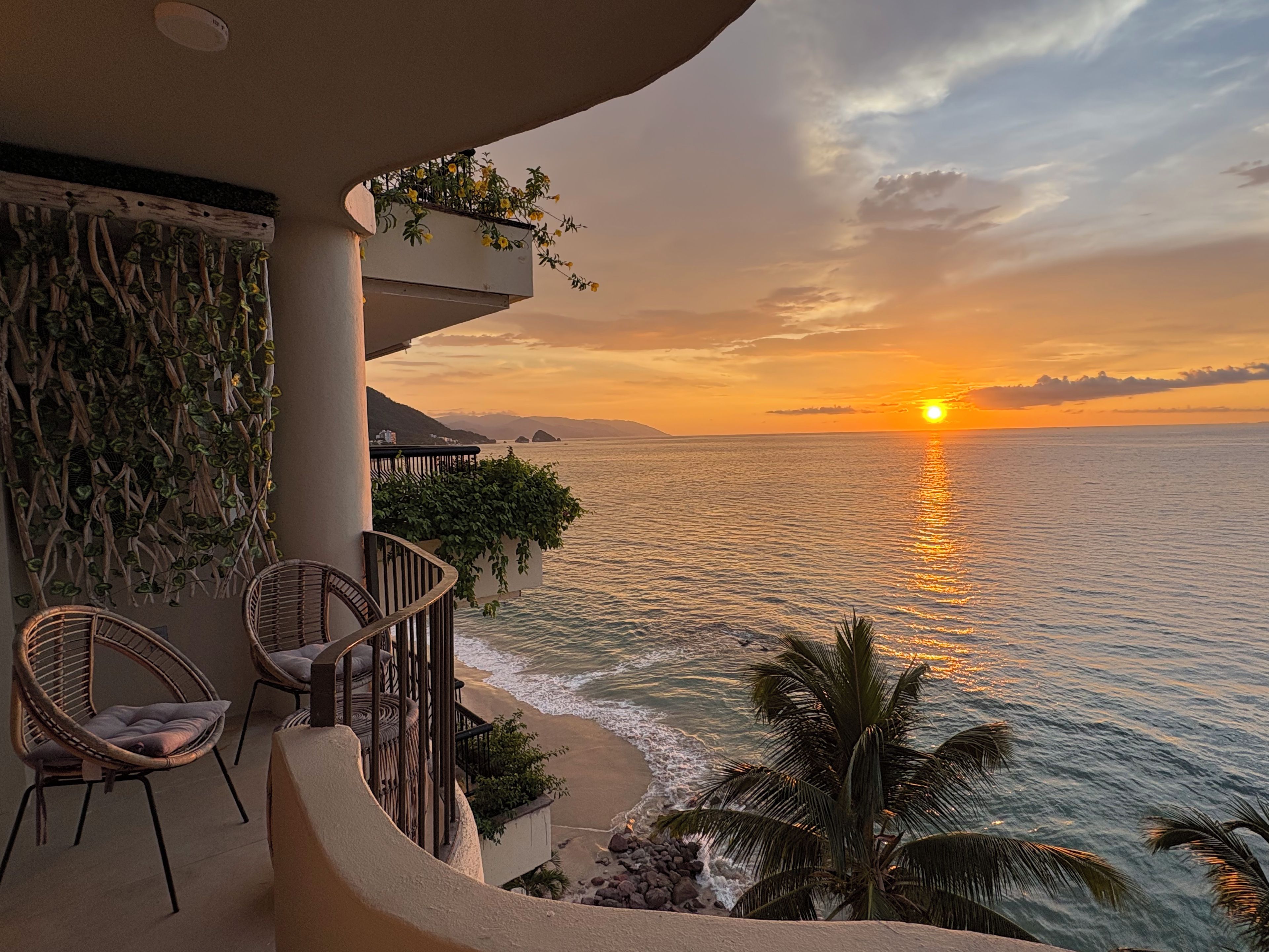 Un balcon avec des fauteuils en osier, une petite table et une vue sur l’océan au coucher du soleil.