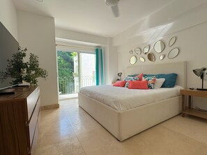 Ein Schlafzimmer mit Bett, Fernseher, Pflanze und Balkon mit Blick auf Grünflächen.