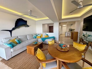 A modern living room with a sofa, coffee table, and dining table. There is a ceiling fan, recessed lighting, and a wall-mounted decorative piece. - Beachfront Boutique Condo - Magical Sunset (Puerto Vallarta)