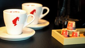 Två vita muggar med röda logotyper, en kaffebryggare och ett bricka med kaffekapslar.