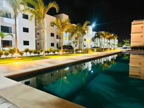 Una piscina amb el reflex d'edificis i palmeres de nit.