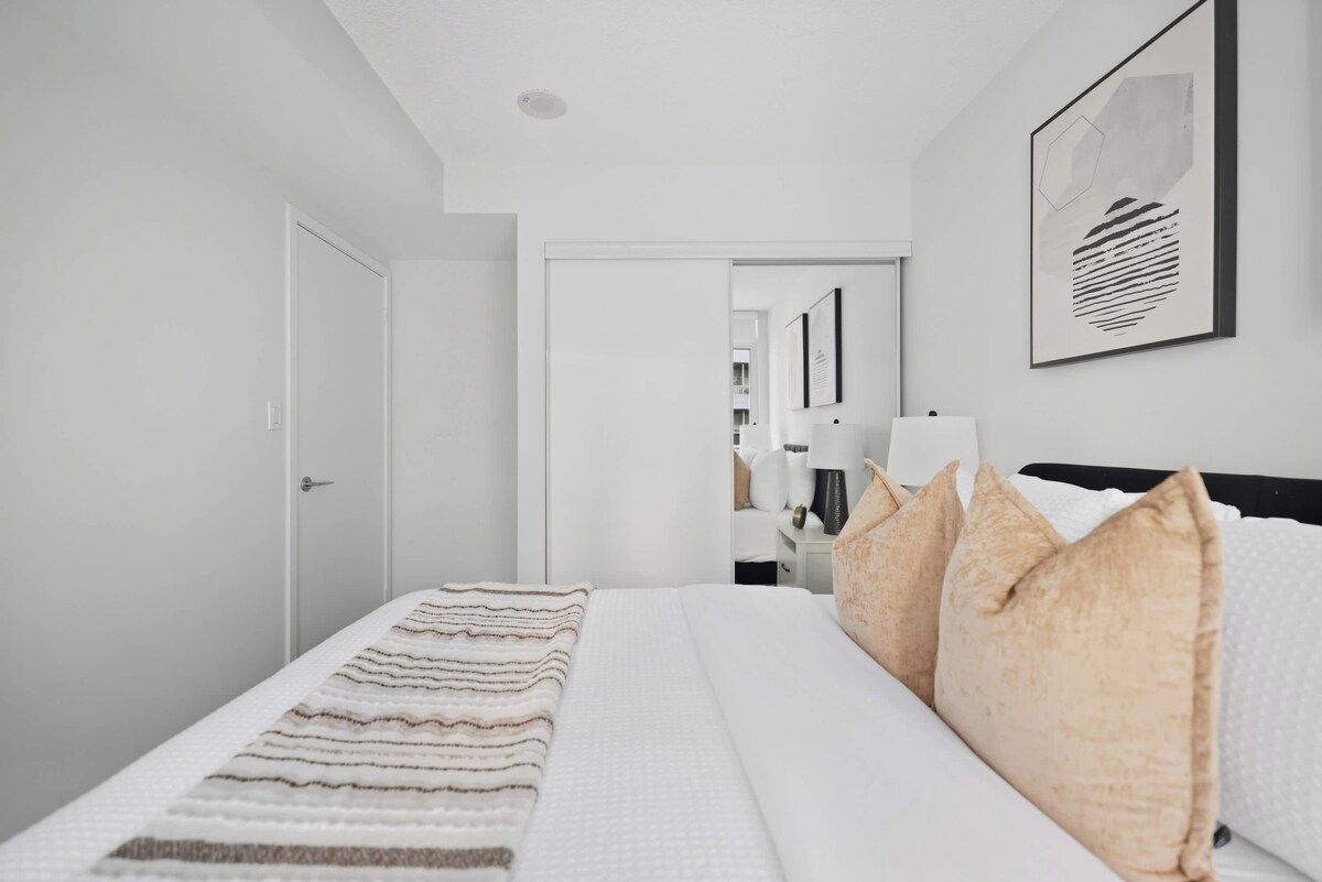 Una habitación de hotel con una cama, almohadas, una mesita de noche, una lámpara y una obra de arte enmarcada en la pared.