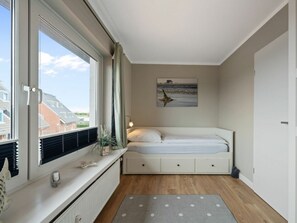 A bedroom with a bed, a window with blinds, a radiator, a wall-mounted artwork, and a small plant on the windowsill. - Utholm 4a - Big Breeze (St. Peter-Ording)
