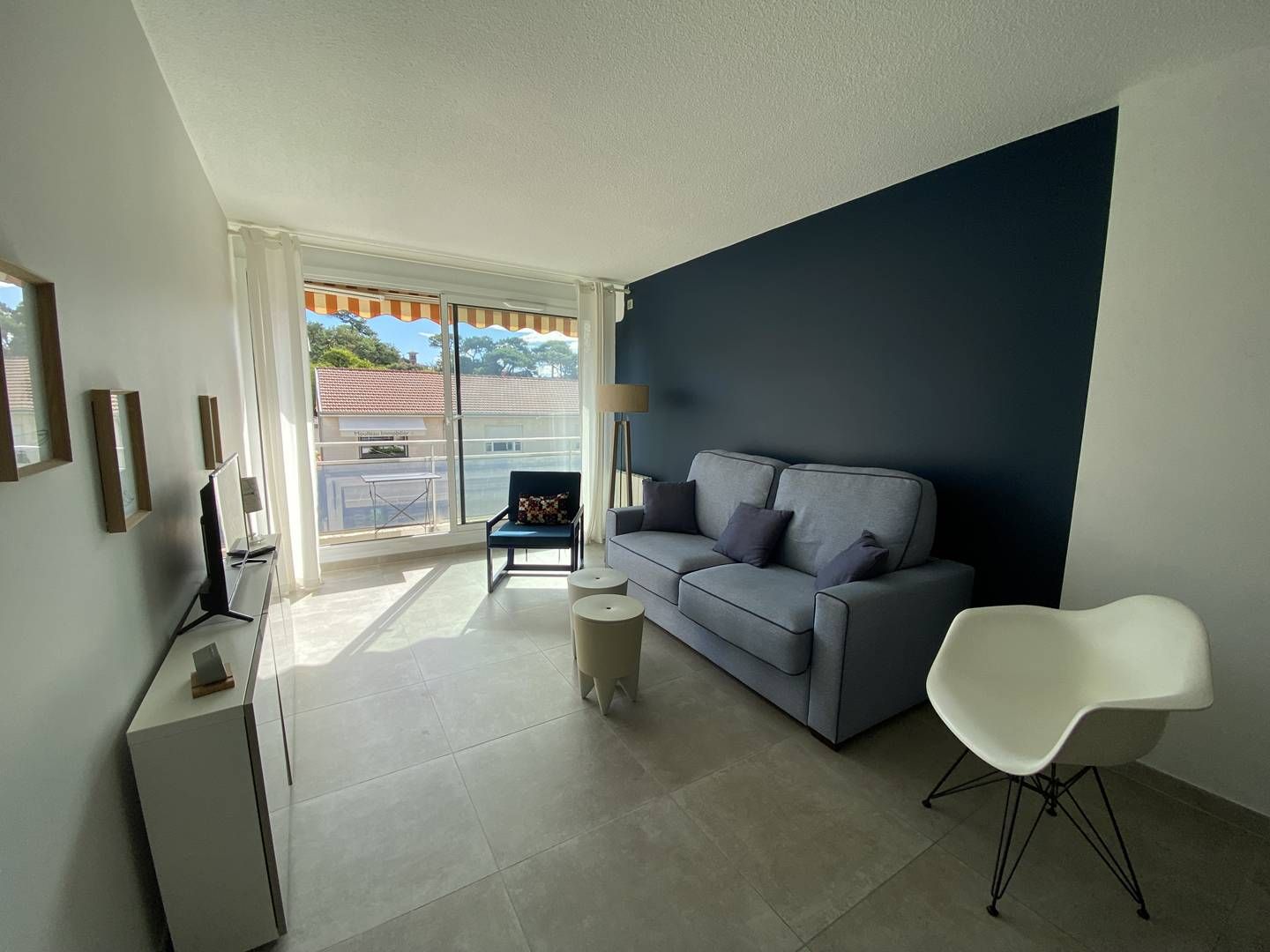 A modern living room with a grey sofa, a white chair, and a small round table. There is a balcony with a view of buildings and trees.