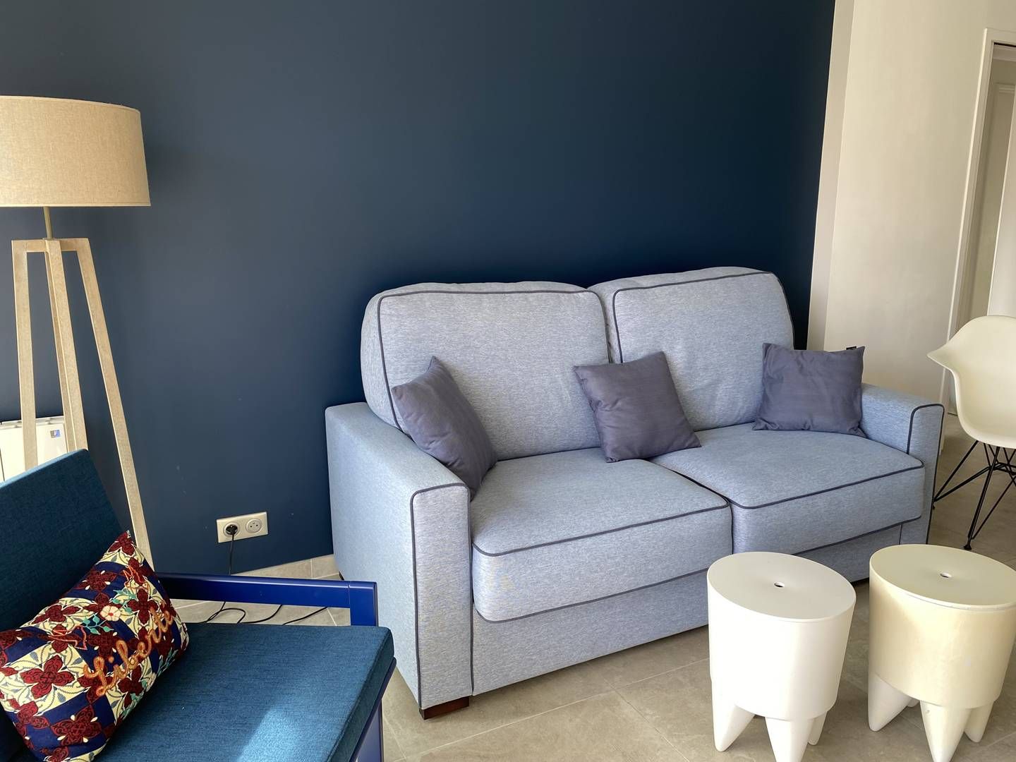 Una sala de estar con un sofá gris, dos mesitas blancas, un sillón azul y una lámpara de pie.