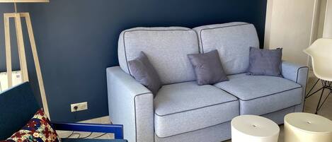 Una sala de estar con un sofá gris, dos mesitas blancas, un sillón azul y una lámpara de pie.