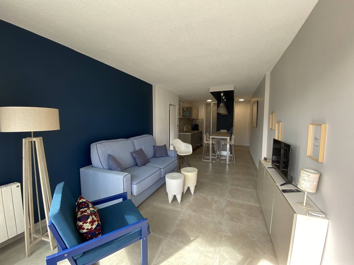 A modern living room with a sofa, a small table, and a chair. There is a kitchen area in the background with a dining table and chairs.