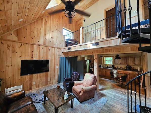 A spacious living area with a wooden interior, a television mounted on the wall, a leather sofa, a coffee table, and a staircase leading to an upper level.
