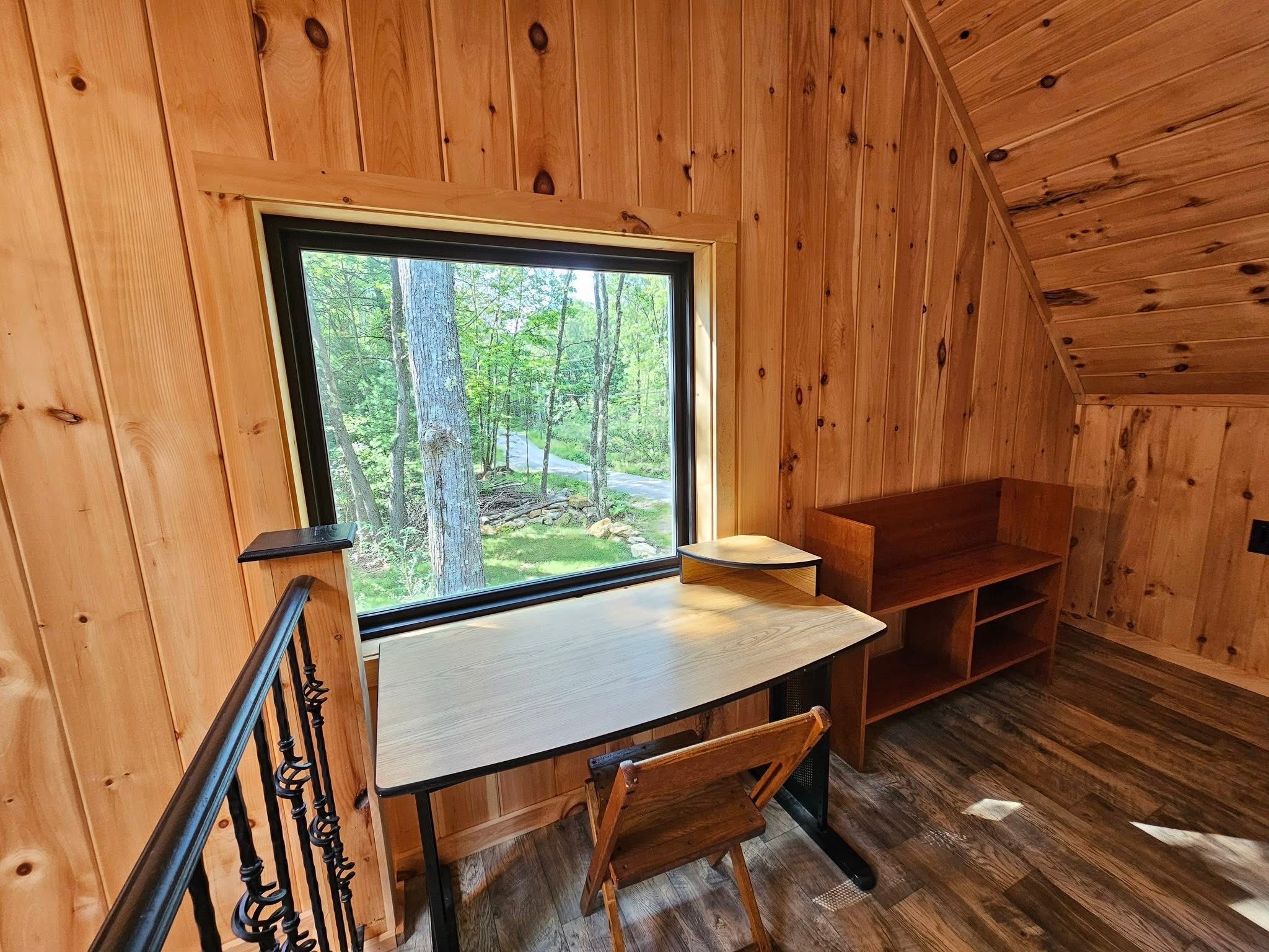 A cabin interior with a wooden desk, chair, and a large window overlooking a forest.