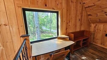 A cabin interior with a wooden desk, chair, and a large window overlooking a forest.