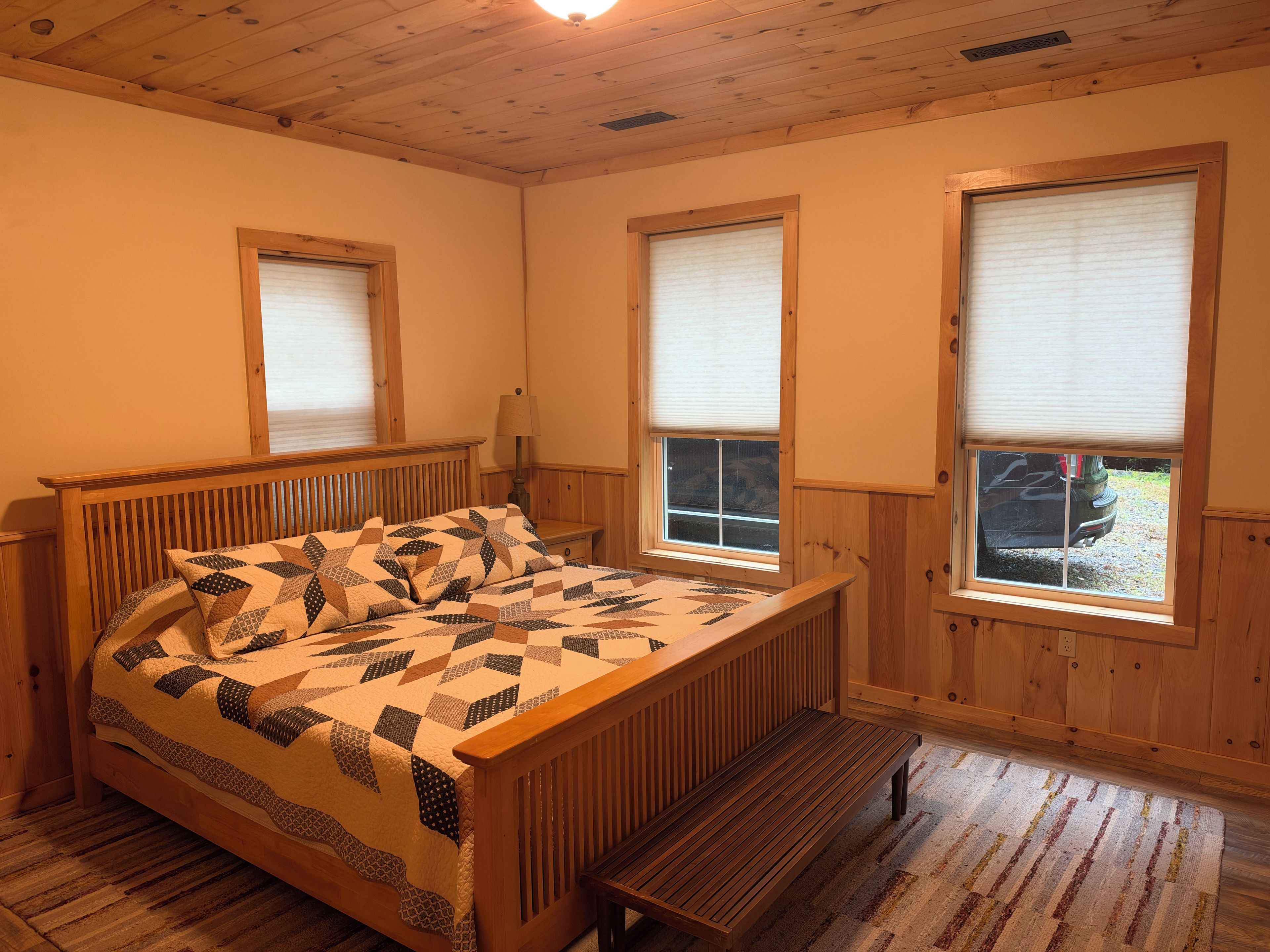 A bedroom with a wooden bed, two windows with blinds, a bench, and a lamp.