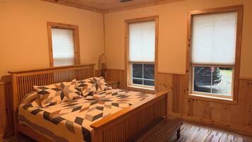 A bedroom with a wooden bed, two windows with blinds, a bench, and a lamp.